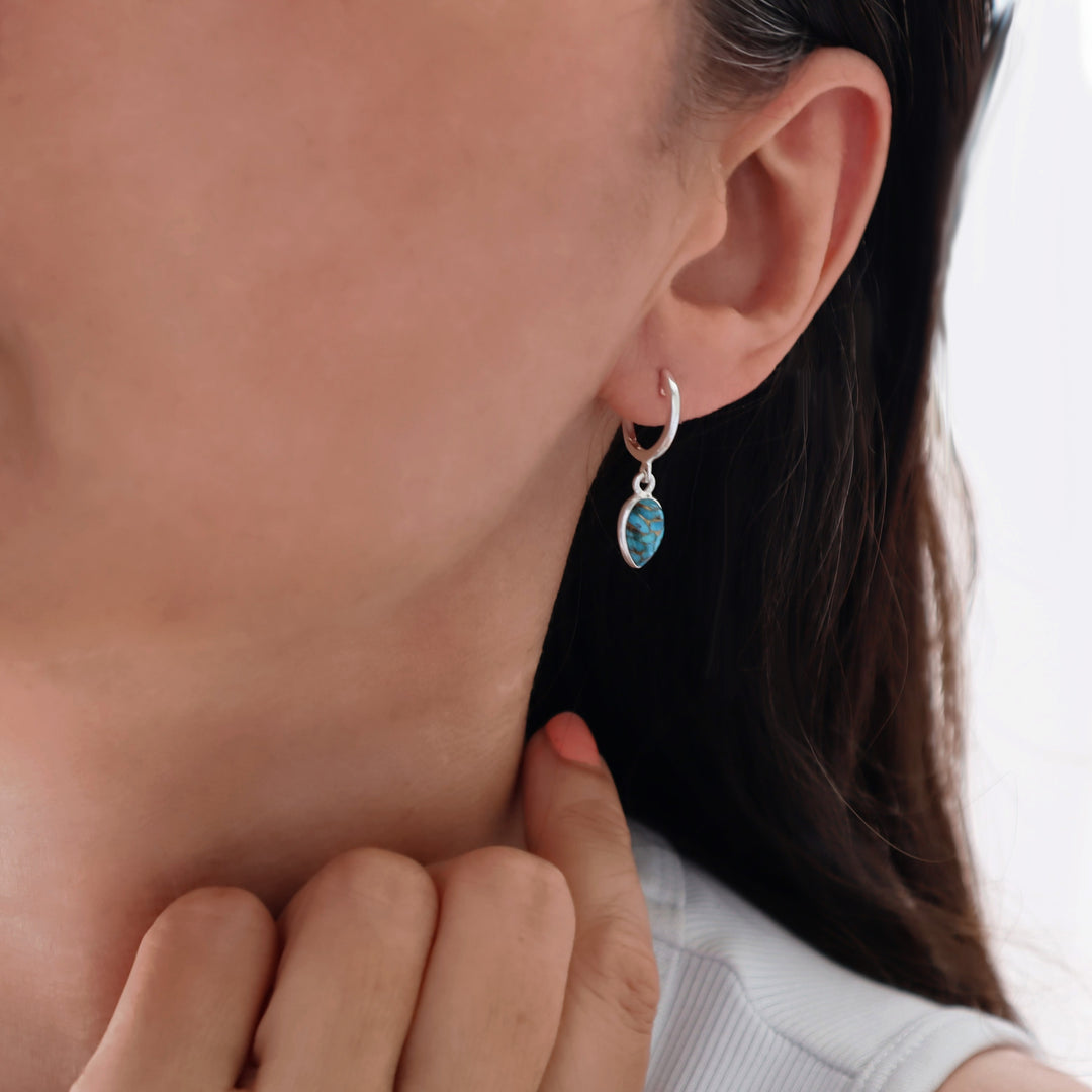 Close up of a woman's ear wearing a 925 siver huggie hoop earring with a dangling blue mojave turquoise gemstone dangling drop. 