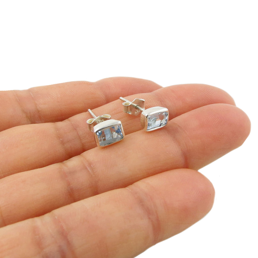 A pair of small silver stud earrings with rectangular baguette cut blue topaz December birthstone gemstones, shown resting on a person's fingers against a white background.