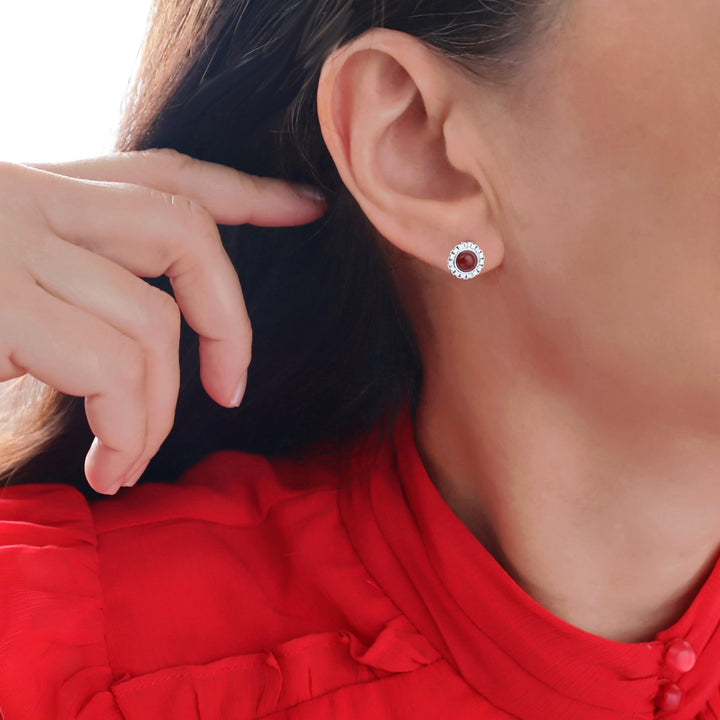 A close-up of a woman wearing a small circular stud earring with a sterling 925 silver halo and a dark red carnelian gemstone centre, shown against her ear and neck with a red top visible.