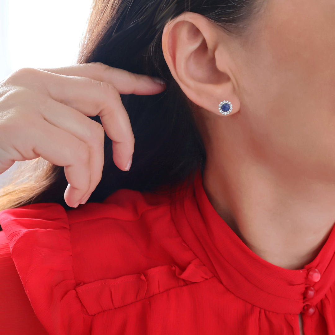 A close-up of a woman wearing a small round 925 silver stud earring with a deep blue lapis lazuli centre and a halo of silver beads, shown against her ear and neck. She is wearing a bright red blouse.
