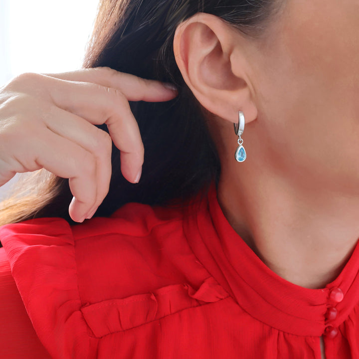 A close-up of a woman wearing a sterling 925 silver huggie hoop earring with a teardrop-shaped light blue aquamarine gemstone, paired with a bright red top.