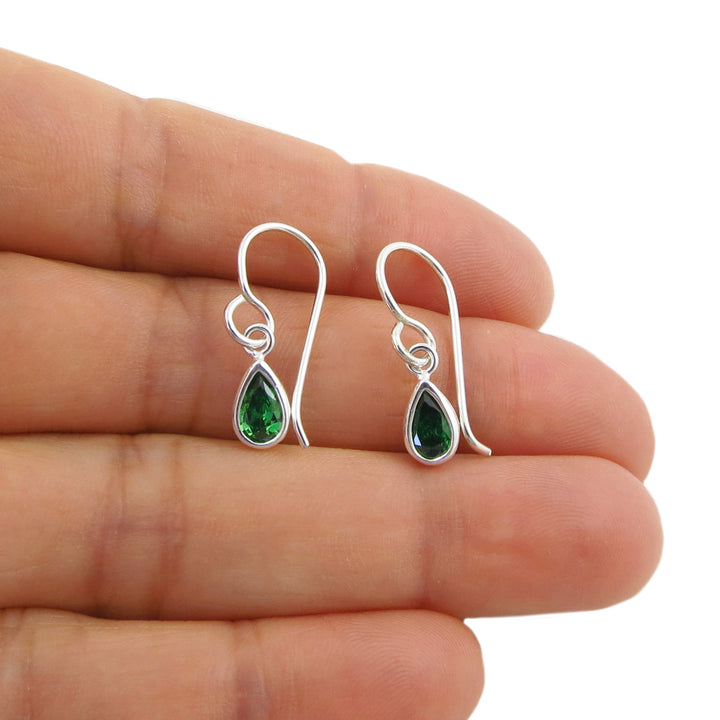 A pair of silver drop earrings with teardrop-shaped green emerald gemstones, shown resting on a person's fingers against a white background.