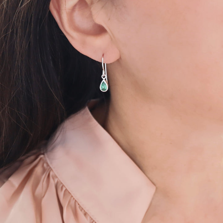 A close-up of a woman's ear wearing a silver drop earring with a small teardrop-shaped green emerald gemstone, paired with a light pink satin blouse.