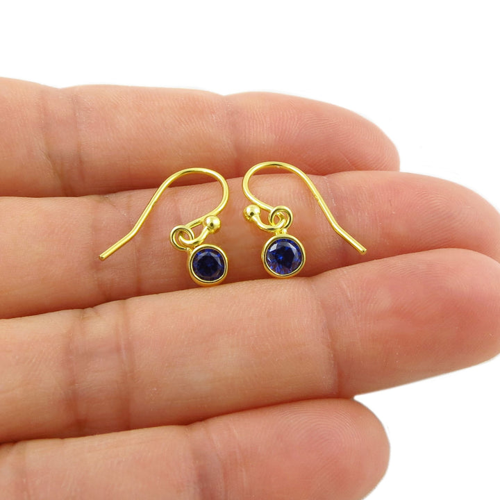 A pair of yellow gold drop earrings with round dark blue sapphire September birthstone gemstones, shown resting on a person's fingers against a white background.