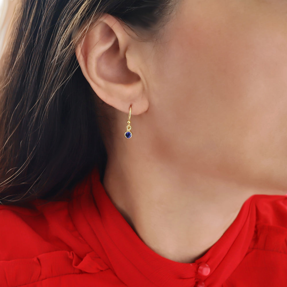A close-up of a woman's ear wearing a delicate yellow gold dangle earring with a small round blue sapphire gemstone, paired with a bright red top.