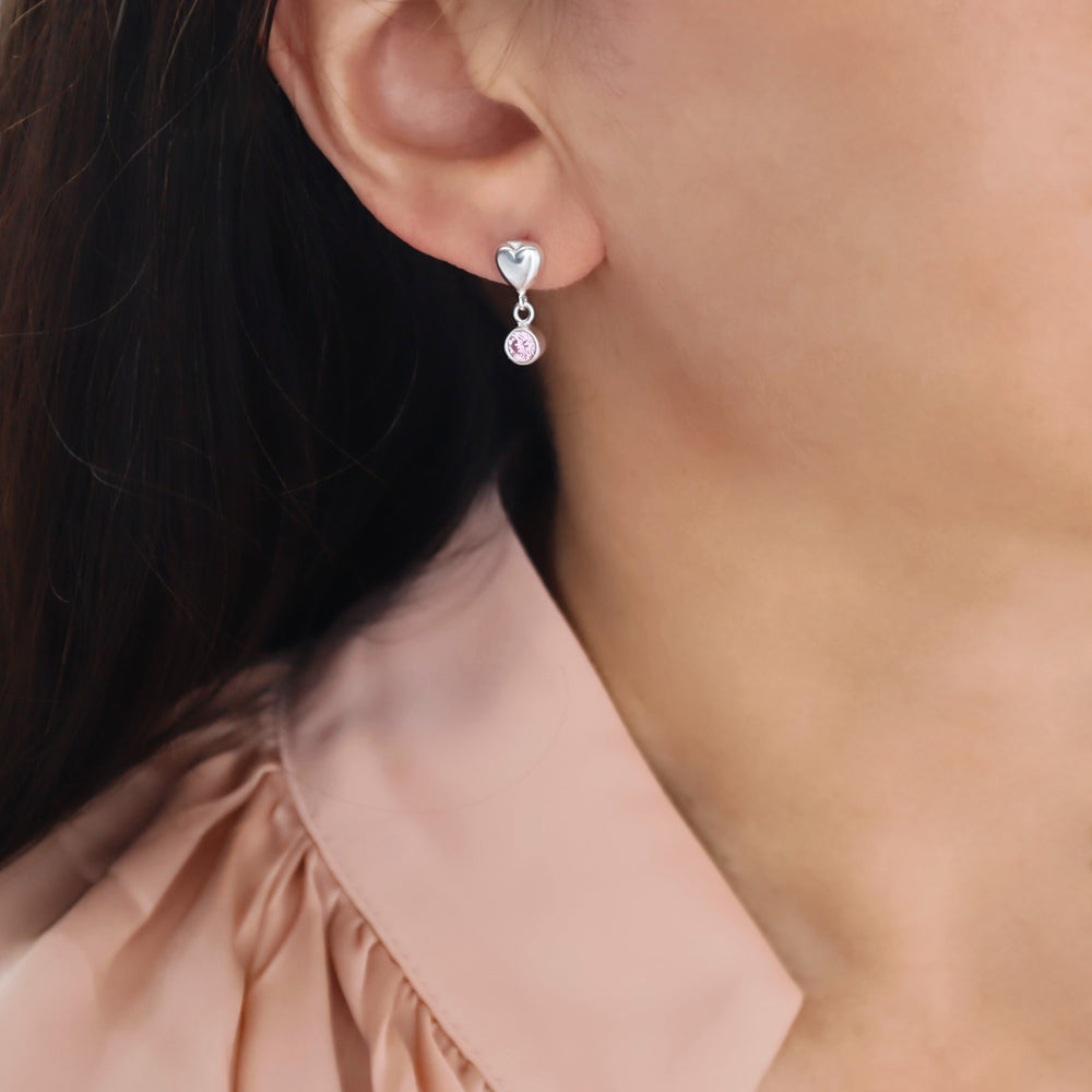 A close-up of a woman's ear wearing a silver heart-shaped stud earring with a dangling pink alexandrite gemstone, paired with a light pink blouse.