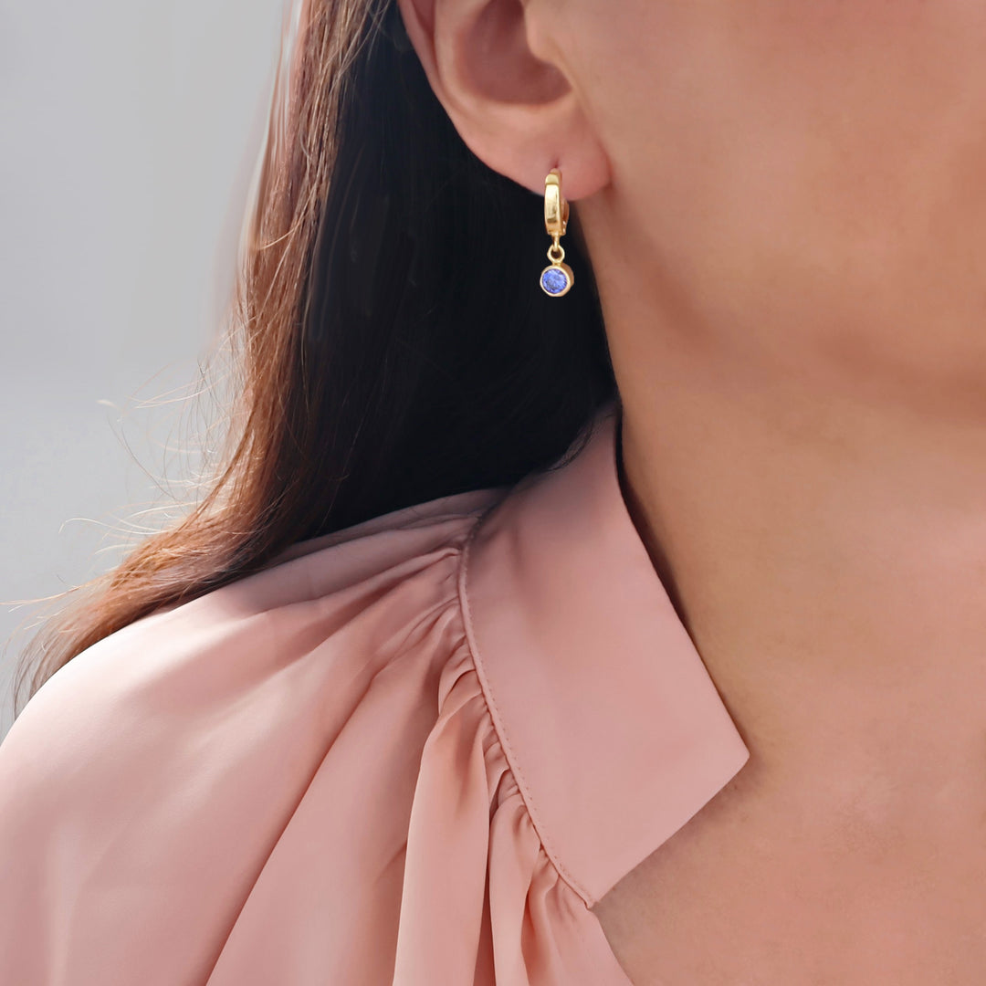 A close-up of a model wearing a silver hoop earring with a small, round blue sapphire September birthstone gemstone, paired with a dusty pink blouse.