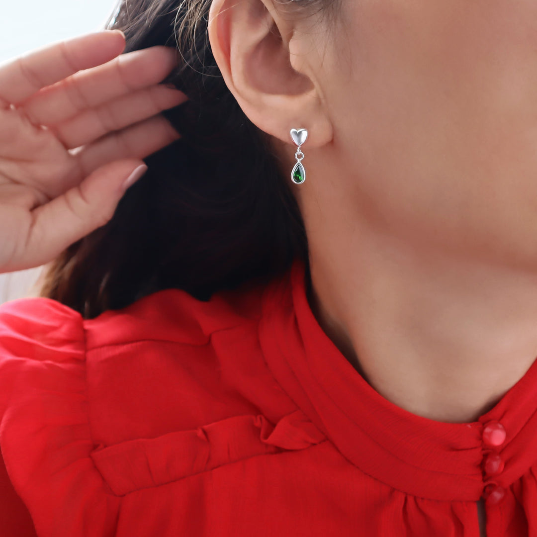 A close-up of a woman wearing a sterling 925 silver heart-shaped drop earring with a green emerald teardrop gemstone, paired with a bright red blouse.