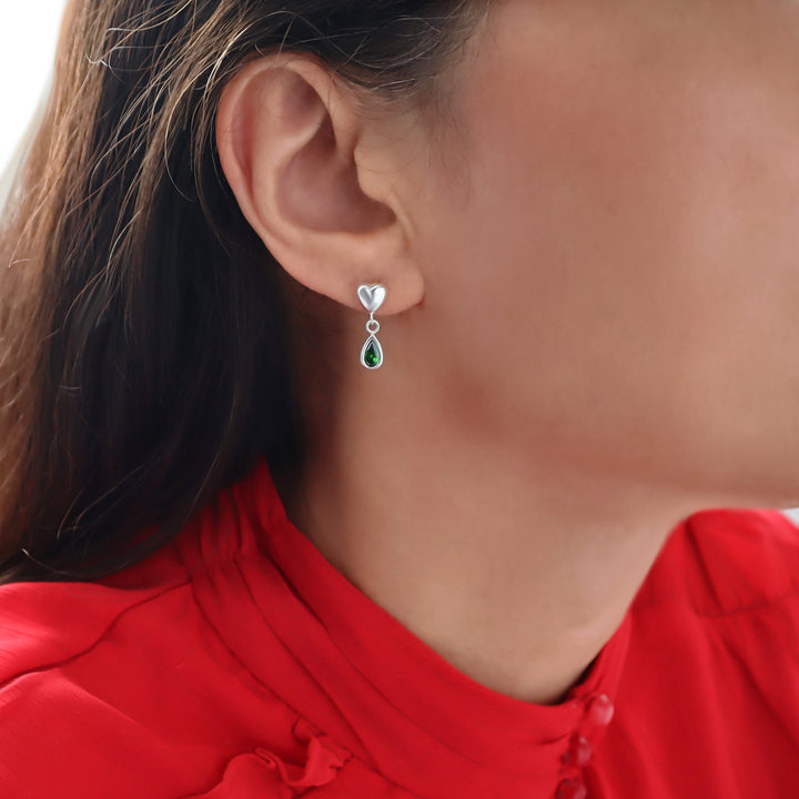 A close-up of a woman's ear wearing a silver dangle earring with a small heart-shaped top and a teardrop green emerald stone drop, paired with a bright red top.