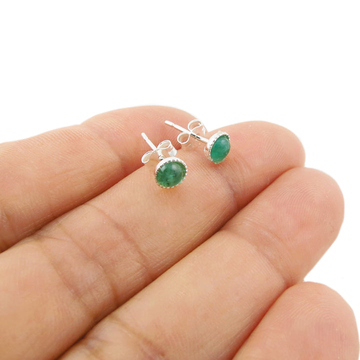 A pair of round natural green emerald gemstone stud earrings in silver settings, shown resting on a person's fingers against a plain white background.