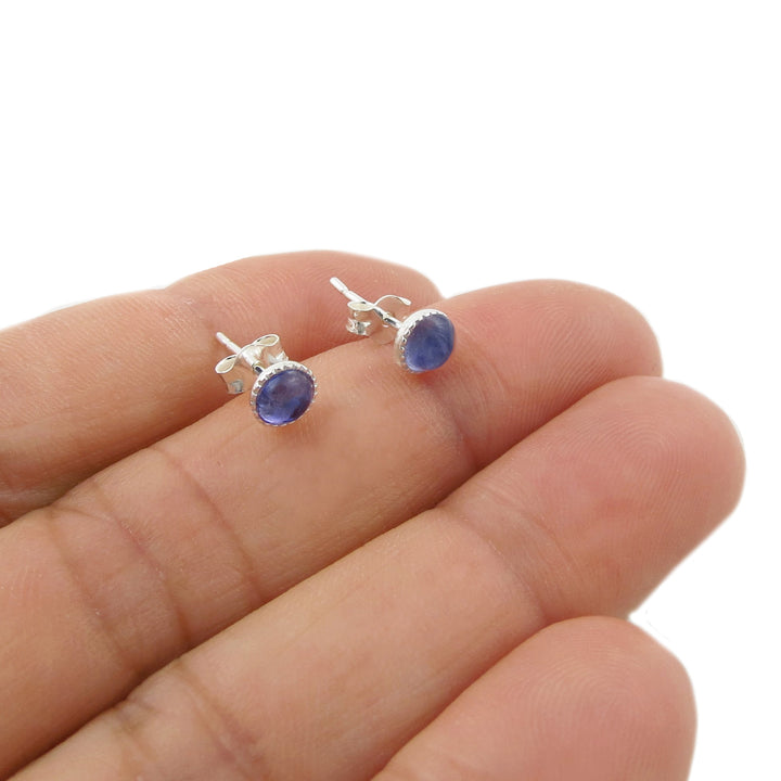 A pair of round blue tanzanite natural gemstone stud earrings with 925 silver settings, shown resting on a person's fingers against a white background.