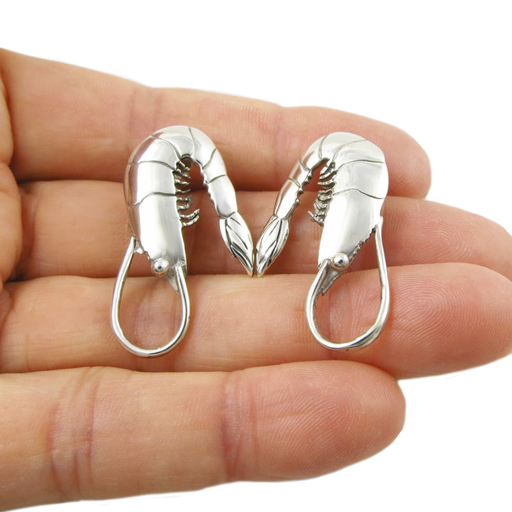 Silver prawn shrimp earrings held in a hand on a white background