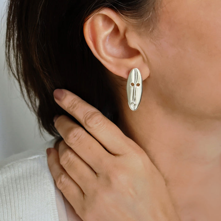 A large tribal mask bohemian 925 sterling silver drop earring worn by a woman with dark hair. 