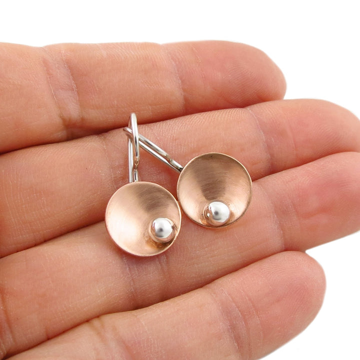 A pair of two tone brushed copper dome stud earrings in a warm copper tone with small 925 silver centres, shown resting on an open hand against a white background.