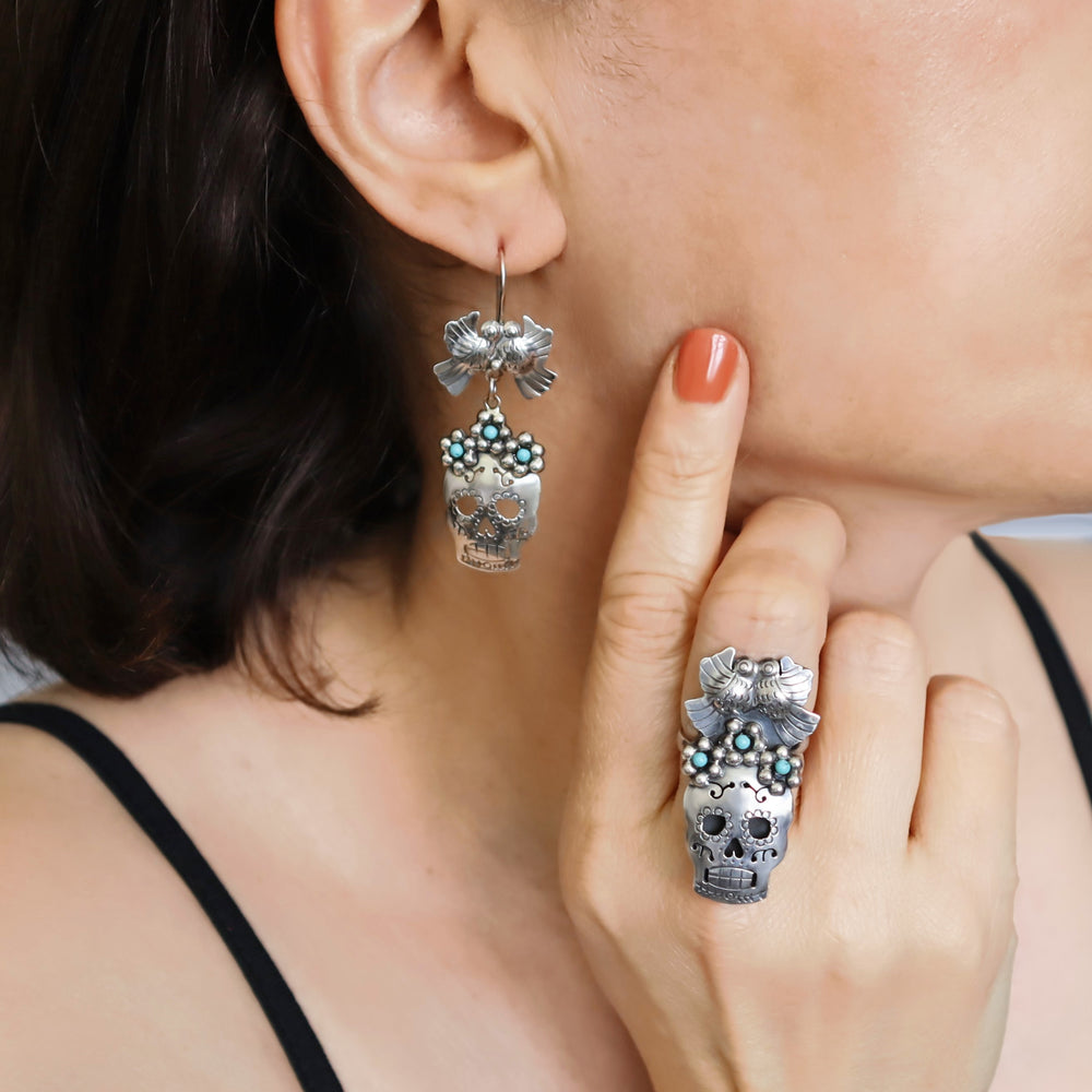 A person wearing silver Taxco sugar skull earrings adorned with flowers and turquoise-coloured beads.