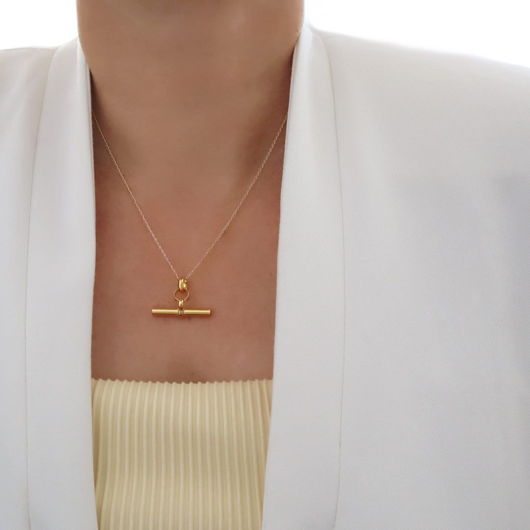 A large gold t-bar pendant and curb chain necklace on a woman wearing a white jacket and yellow top