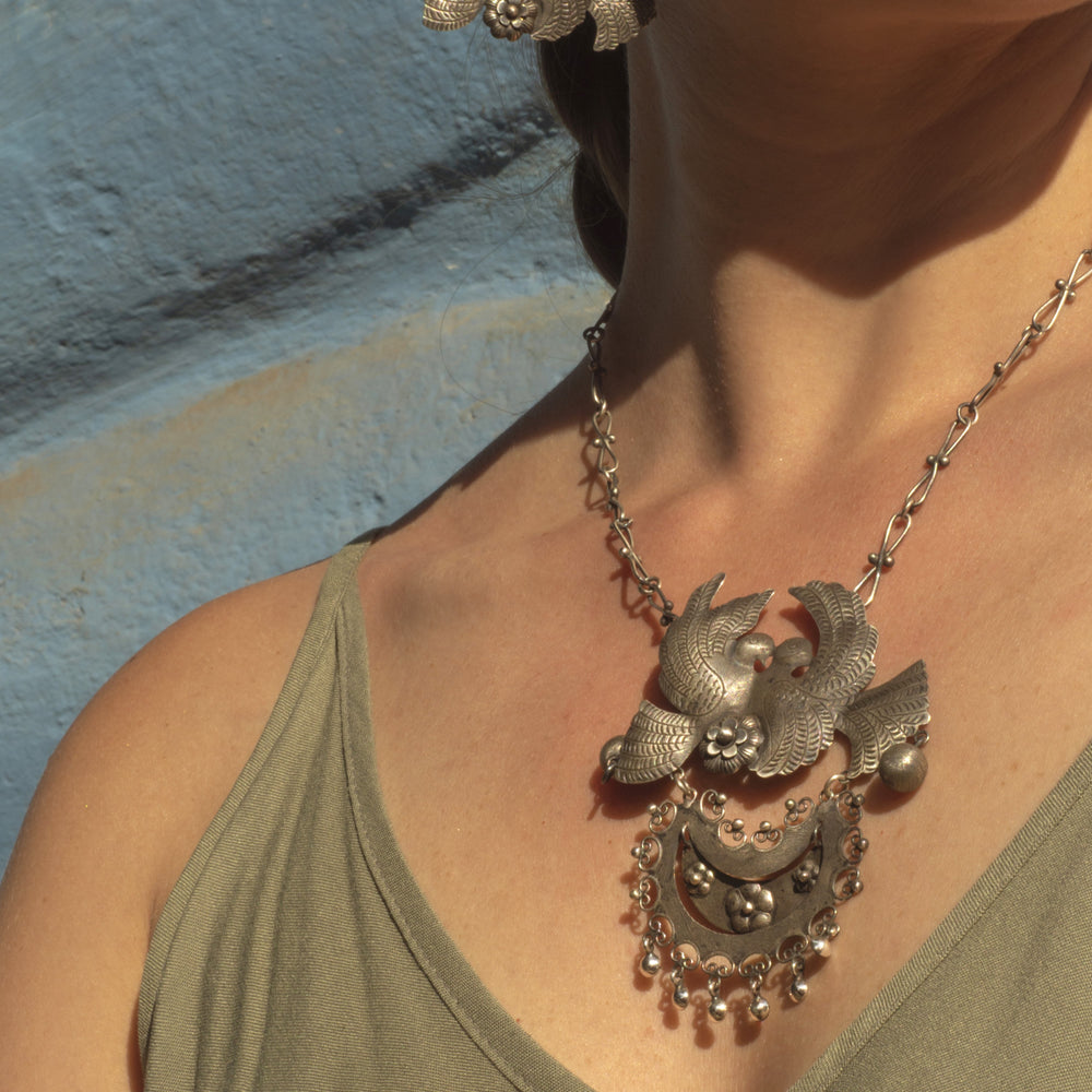 A close-up of a woman wearing a detailed silver pendant necklace featuring two birds and a crescent moon, paired with a green top against a textured blue wall.