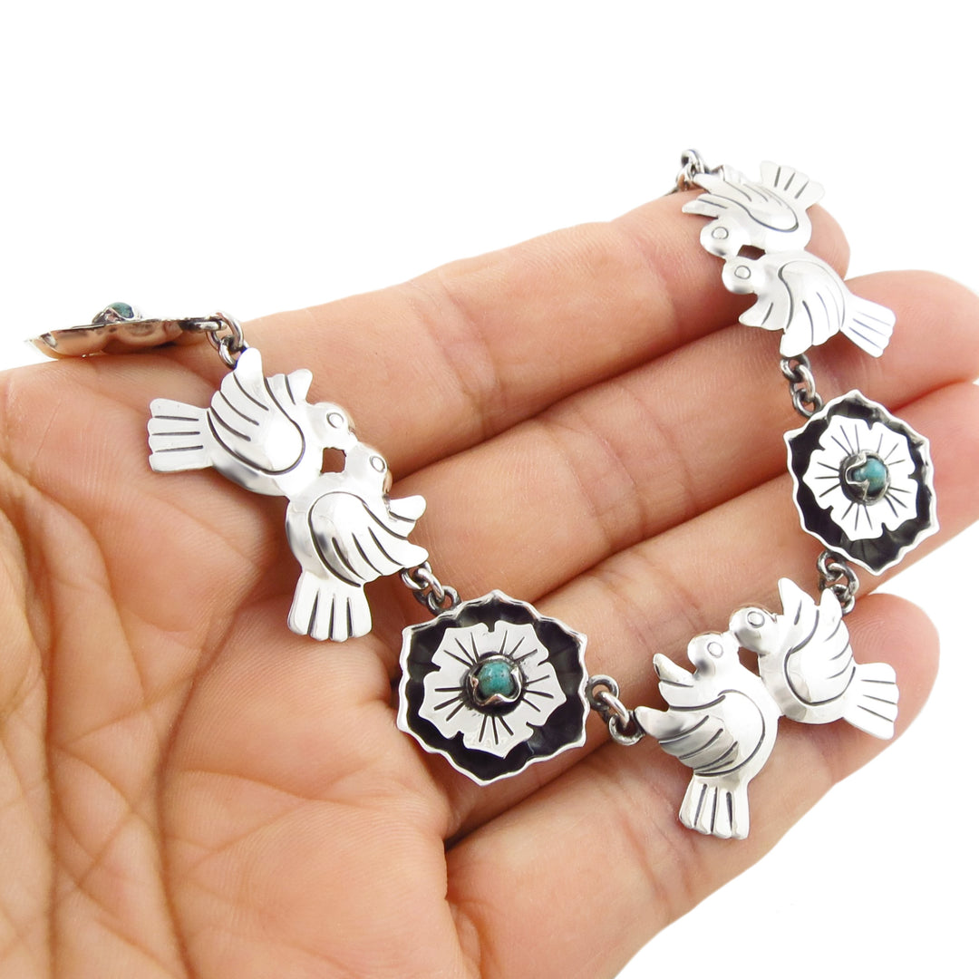 A Mexican sterling 925 silver necklace with bird and floral motifs with turquoise gemstones, held in a person's hand against a white background.