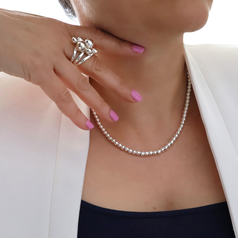 Silver ball bead necklace being worn with a hand holding a silver ball ring, against a white background