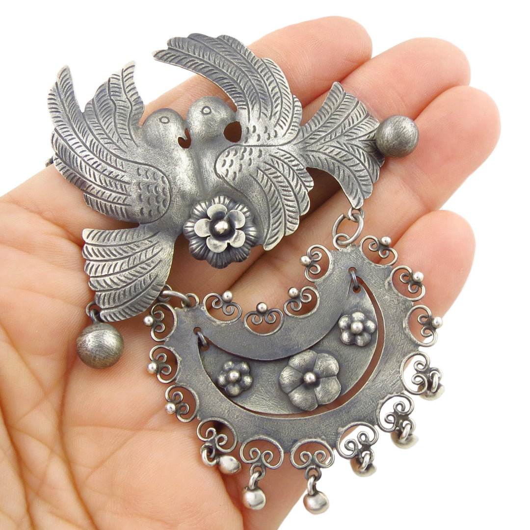An ornate sterling 925 silver pendant and chain necklace featuring two love birds, a crescent moon, floral motifs, and dangling beads, held in a woman's hand against a plain white background.