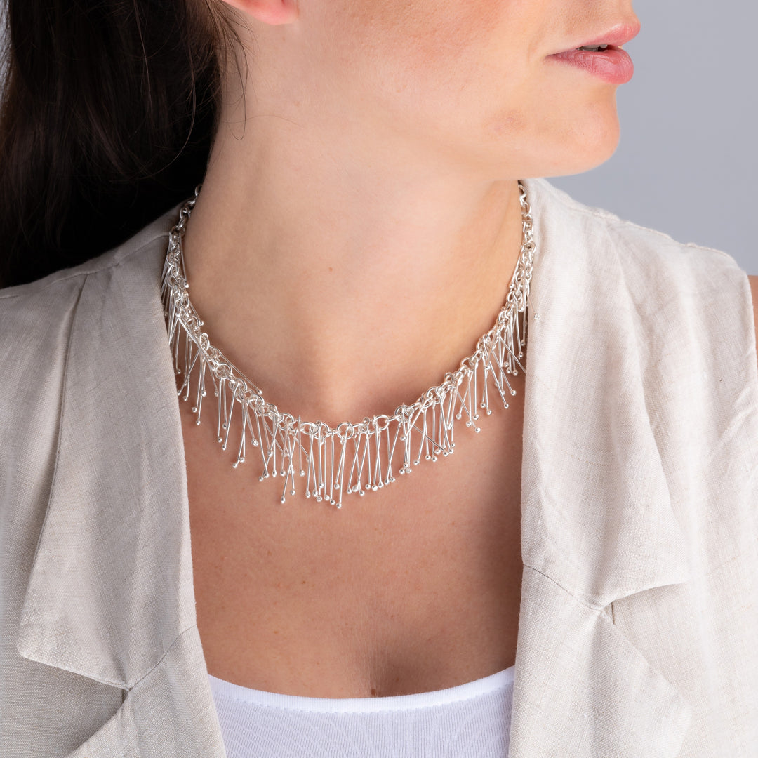 A long bib necklace in UK sterling 925 silver featuring a fringe of dangling stick drops worn by a woman with a white jacekt. 