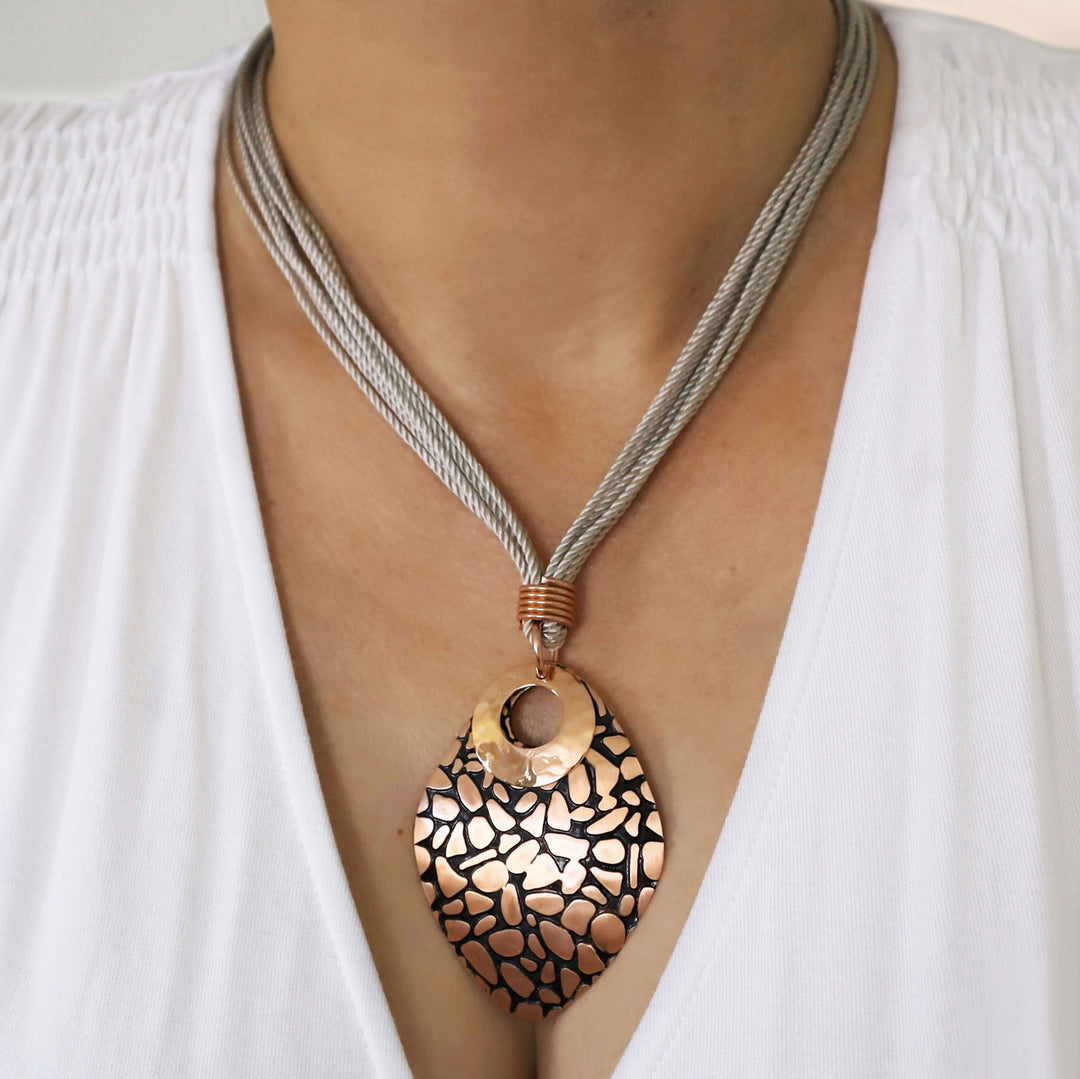 Woman wearing a large animal print copper pendant suspended from a multi-strand gray cord, against a white top.