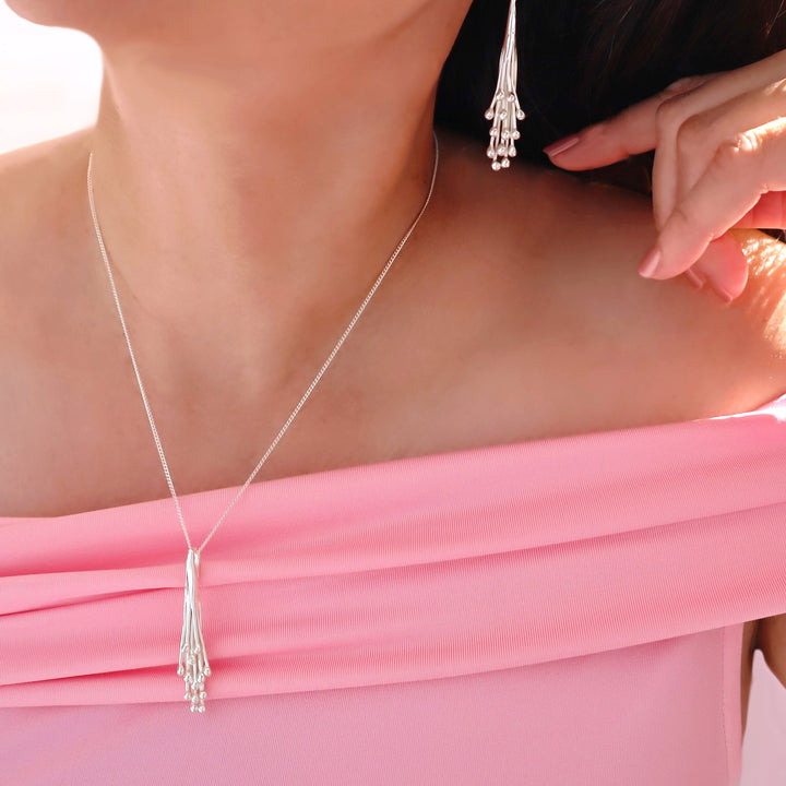 A close-up lifestyle shot of a model wearing a delicate silver necklace with a tassel-like pendant and matching dangling earrings, styled with a pink off-the-shoulder top.