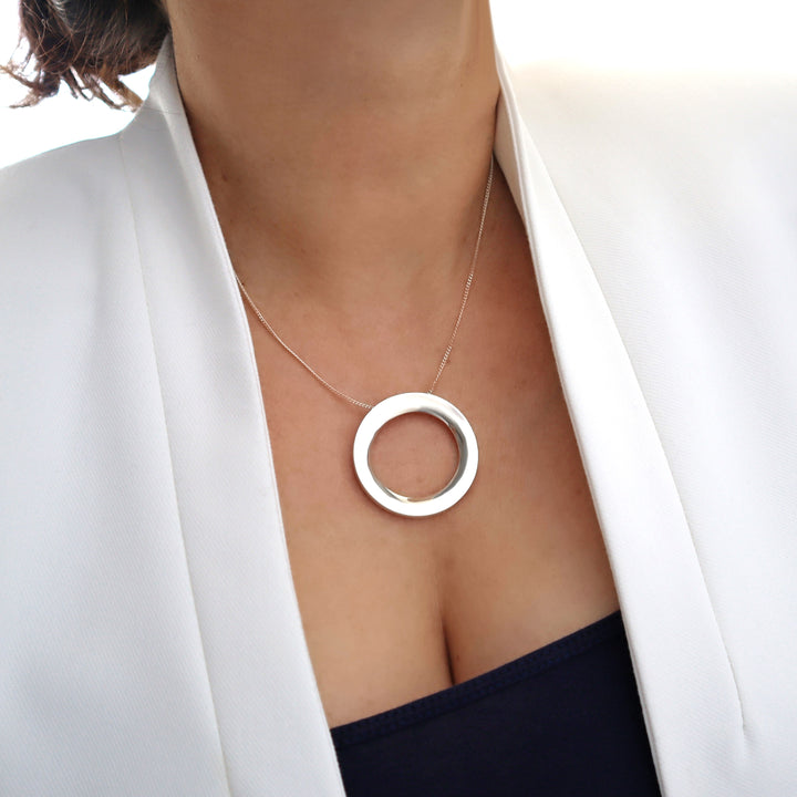 Silver necklace with a circular pendant worn by a person in a white blazer.