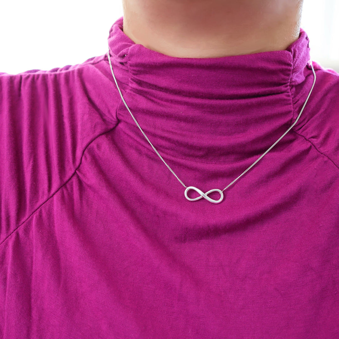 A sterling silver infinity symbol pendant and curb chain necklace worn by a woman in a purple high neck top. 