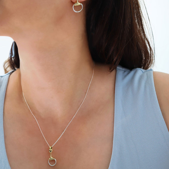 A two tone snaffle horsebit pendant necklace in 925 silver and gold with matching earrings worn by a woman with dark hair 
