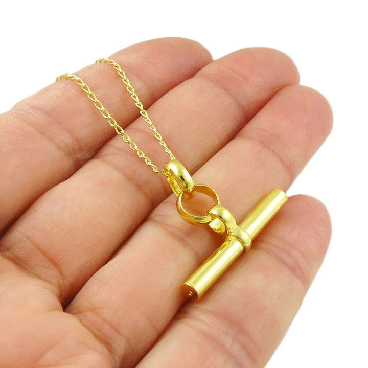 A bright yellow gold large t-bar pendant and curb chain necklace held in a persons hand on a whte background