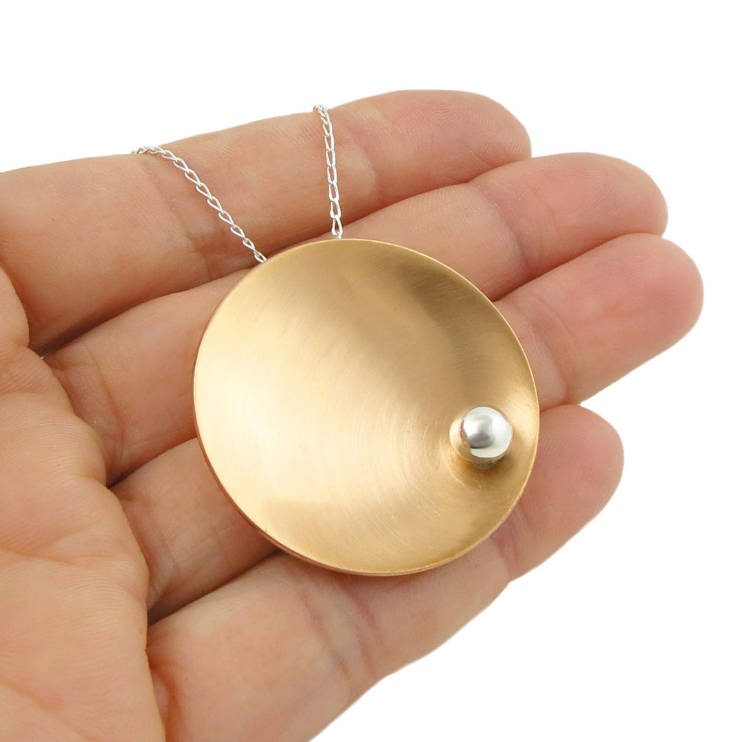 Brushed copper pendant with a small silver ball bead accent hanging on a silver curb chain held in a woman's hand against a white background