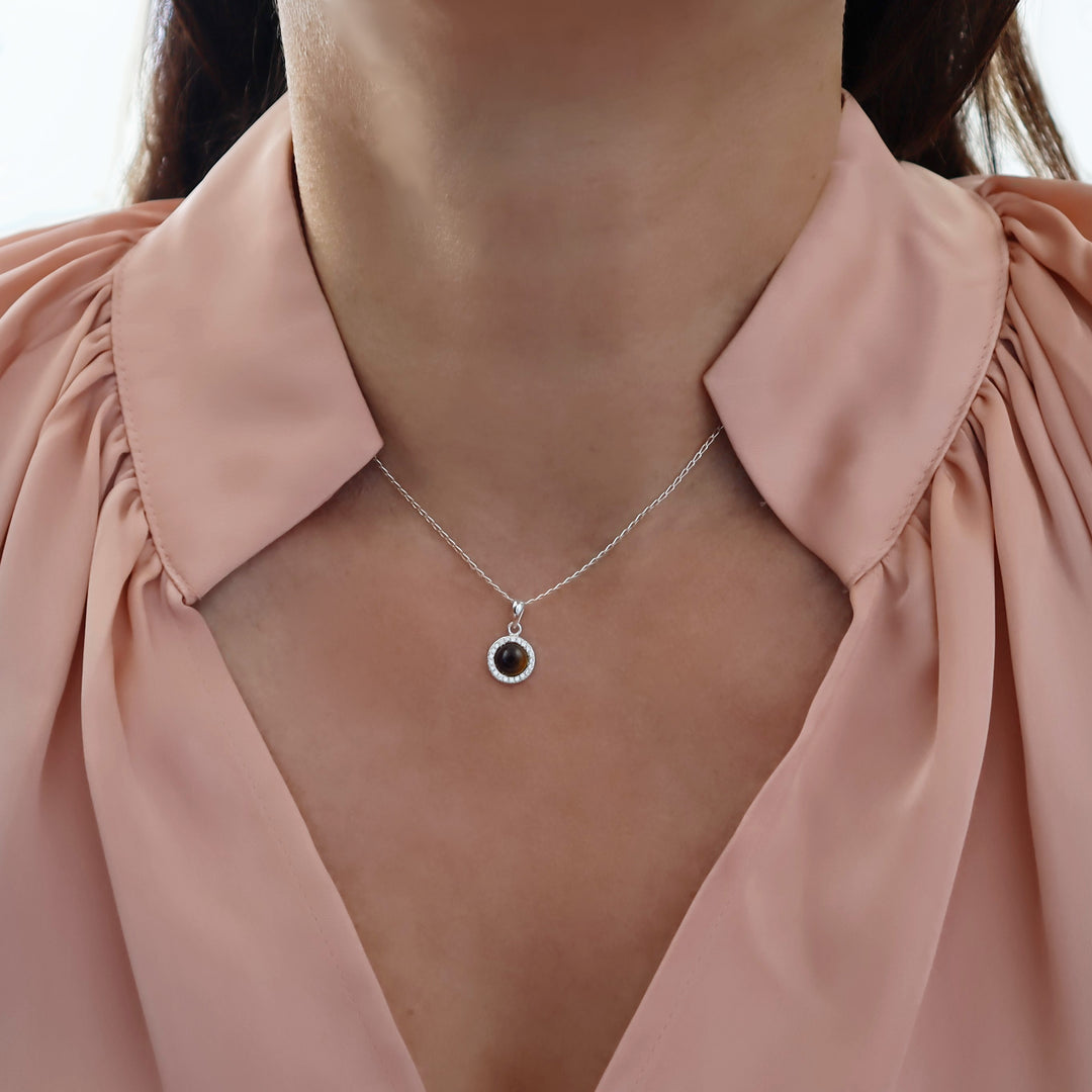A close-up of a model wearing a delicate silver chain necklace with a small circular pendant featuring a dark brown tiger's eye gemstone in the centre, styled with a dusty pink blouse.