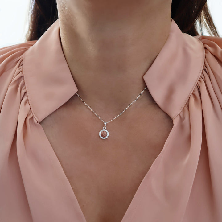 A close-up of a model wearing a delicate silver chain necklace with a circular pendant featuring a pink rhyolite gemstone and a halo of small clear stones, styled with a dusty pink blouse.