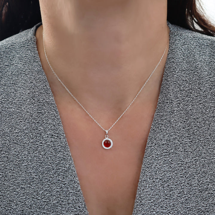 A close-up of a woman wearing a delicate 925 silver chain necklace with a small circular pendant featuring a red carnelian agate gemstone and a halo of clear stones, styled with a gray textured top.