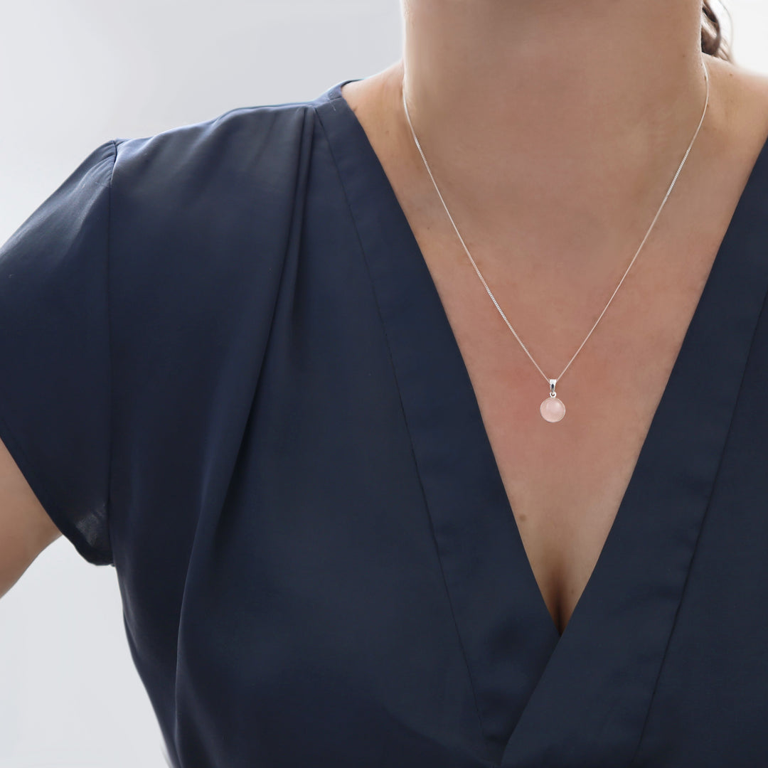 A close-up of a model wearing a delicate silver chain necklace with a small, round pale pink rose quartz gemstone pendant, paired with a navy blue satin top.