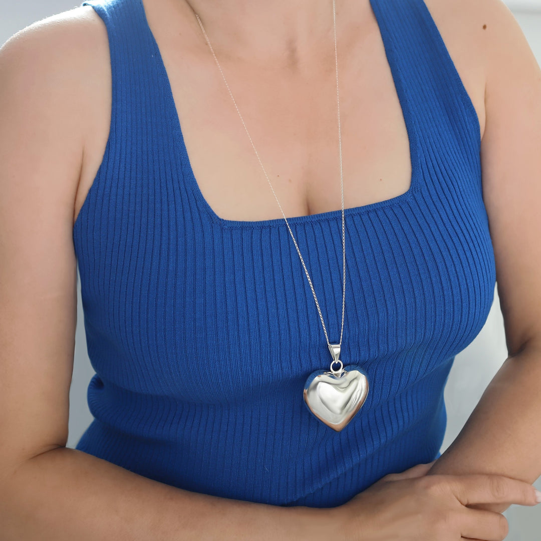 A woman in a blue top wearing a large over-sized sterling silver puffed heart pendant on a long silver curb chain, 