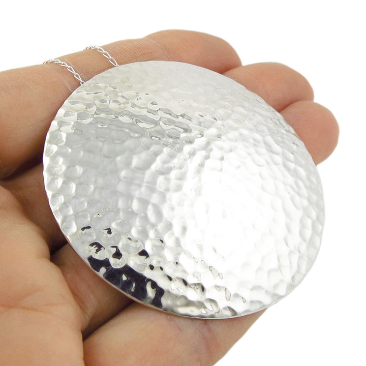 An extra large UK hallmarked sterling 925 silver hammered pendant and chain necklace for women held in a hand against a white background