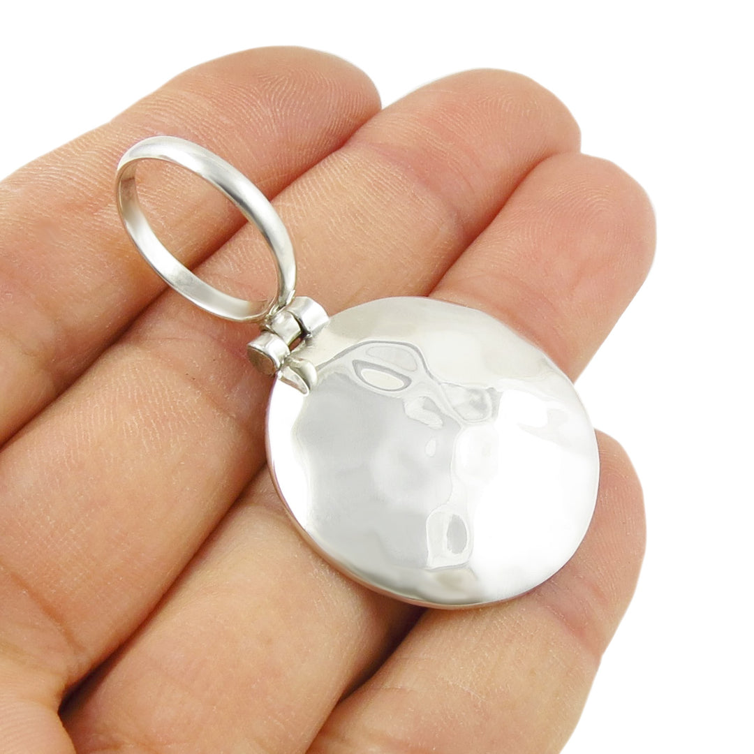 A large silver pendant with a hammered finish and a large bail held in a person's hand. 