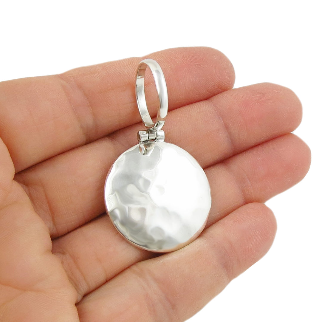 A round hammered pendant in sterling silver with a large over-sized bale held in a person's hand. 