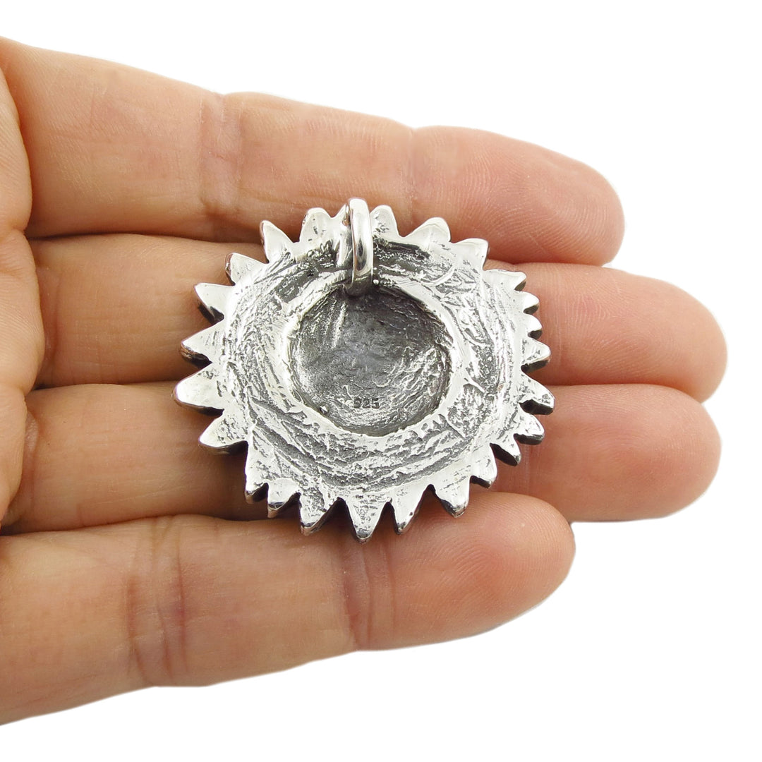 Reverse of a sterling silver sunflower pendant with a textured, hammered finish and a bail at the top, held in the palm of a hand against a white background.