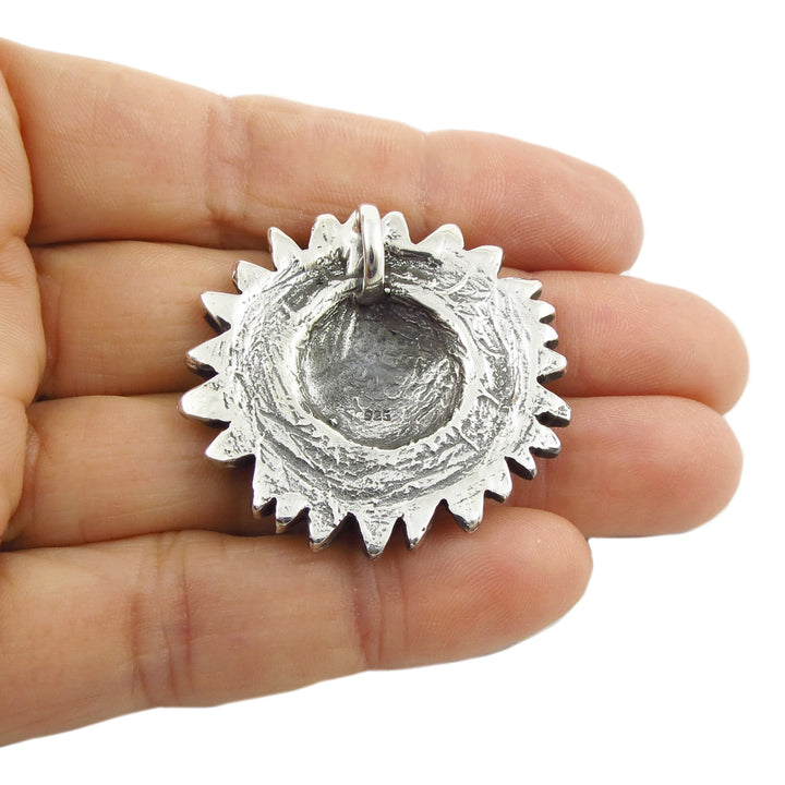 Reverse of a sterling silver sunflower pendant with a textured, hammered finish and a bail at the top, held in the palm of a hand against a white background.