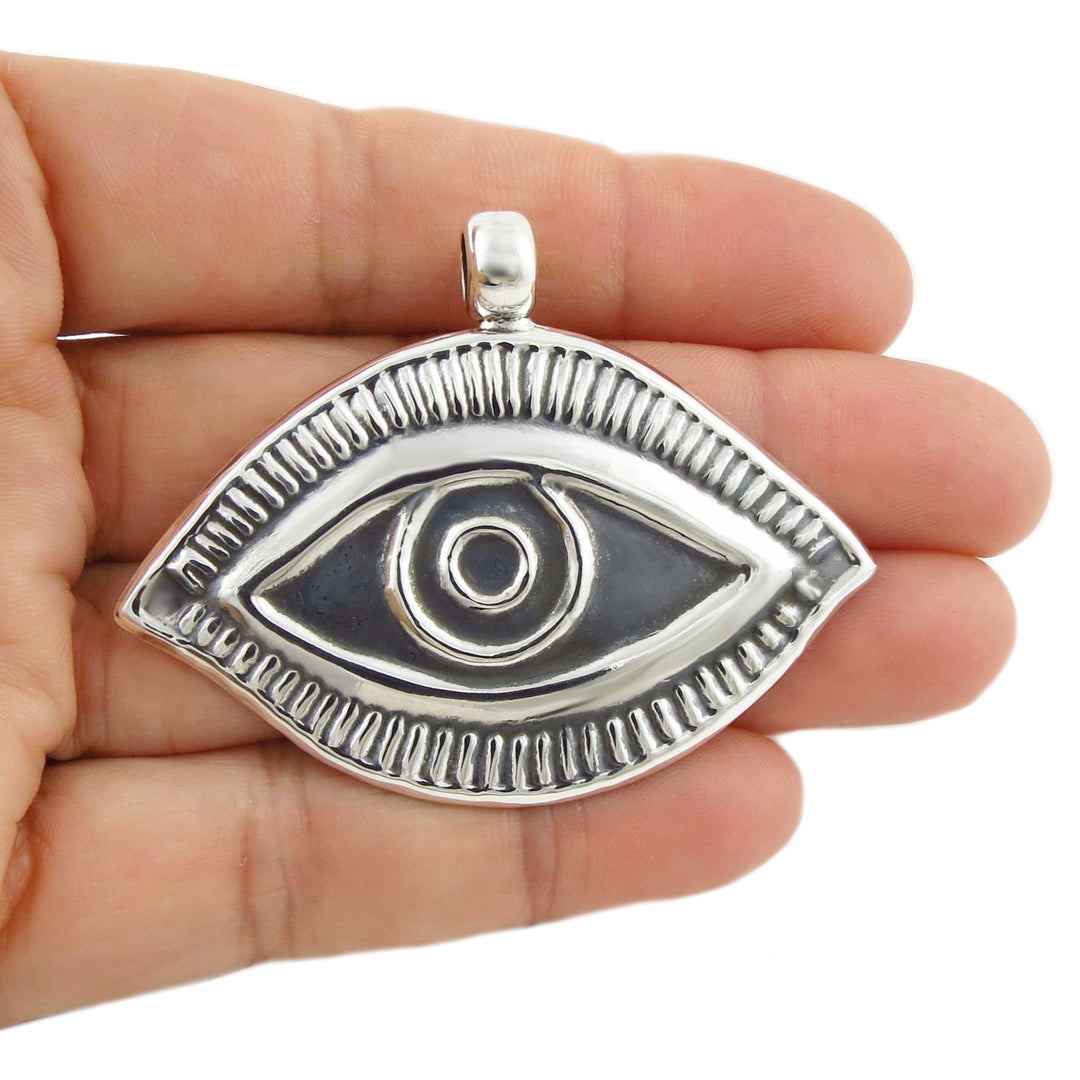 A sterling silver eye-shaped lucky magic eye pendant with a textured border and raised pupil detail, held in the palm of a hand against a white background.