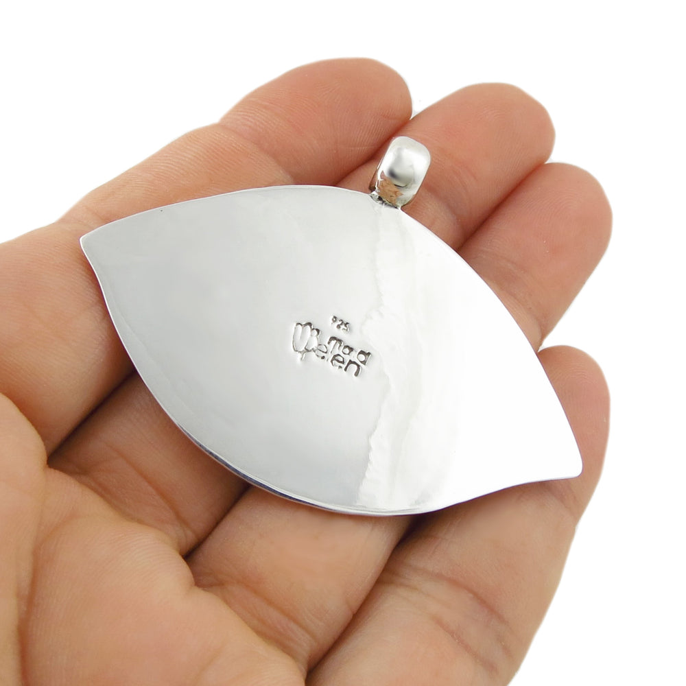 A polished silver pendant shaped like an eye, held in a person's hand. The pendant features a small engraved Matia Belen Nilson logo in the center and a polished silver bail at the top.