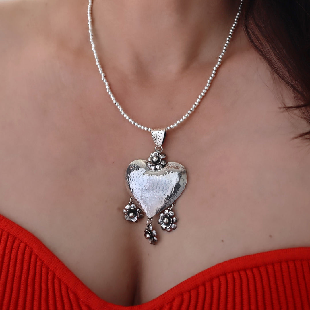 A close-up of a woman wearing a silver heart pendant necklace with a textured finish, a beaded chain, and small dangling floral details. The woman is wearing a bright red ribbed top.
