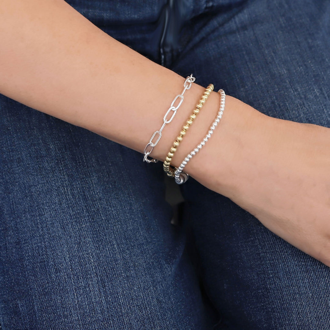 Close-up of a wrist wearing a 925 silver and gold multiple paperclip and ball bracelets in a wrist stack on a blue fabric background