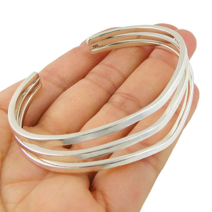 A sterling silver bracelet featuring three cufs that join at the ends held in a person's hand on a white background. 