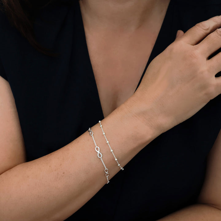 Woman wearing a wrist stakc featuring a curb chain stacking bracleet with figure of eight links and another circle link chain decorated with silver ball beads