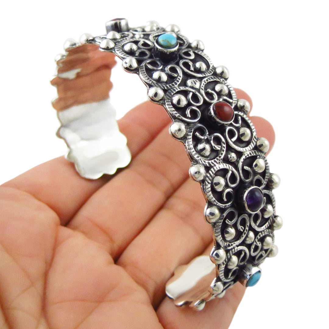 A hand holding a large sterling 925 silver filigree cuff bracelet with turquoise, red, and purple gemstone accents.
