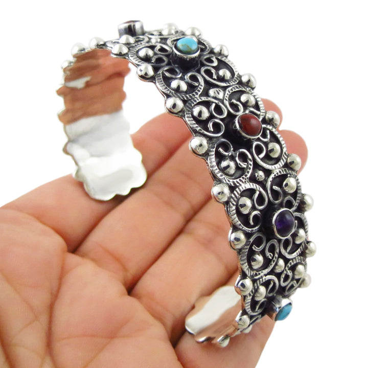 A hand holding a large sterling 925 silver filigree cuff bracelet with turquoise, red, and purple gemstone accents.